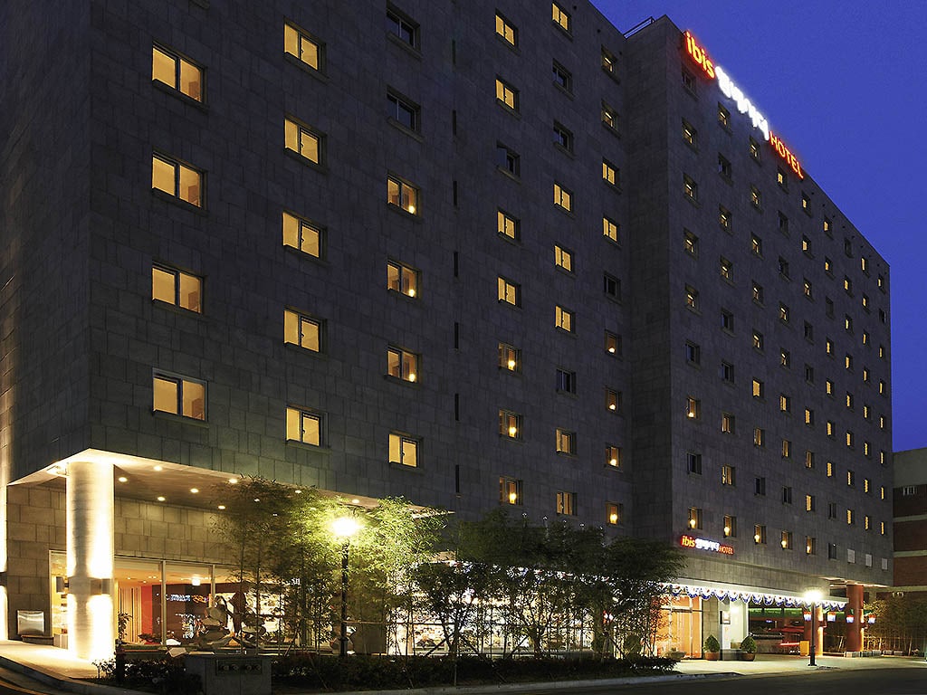 A modern, multi-story hotel building at night with many lit windows, a well-lit entrance, and a canopy above the main door. The hotels name is illuminated in red and orange letters on the rooftop. Trees line the entrance area.