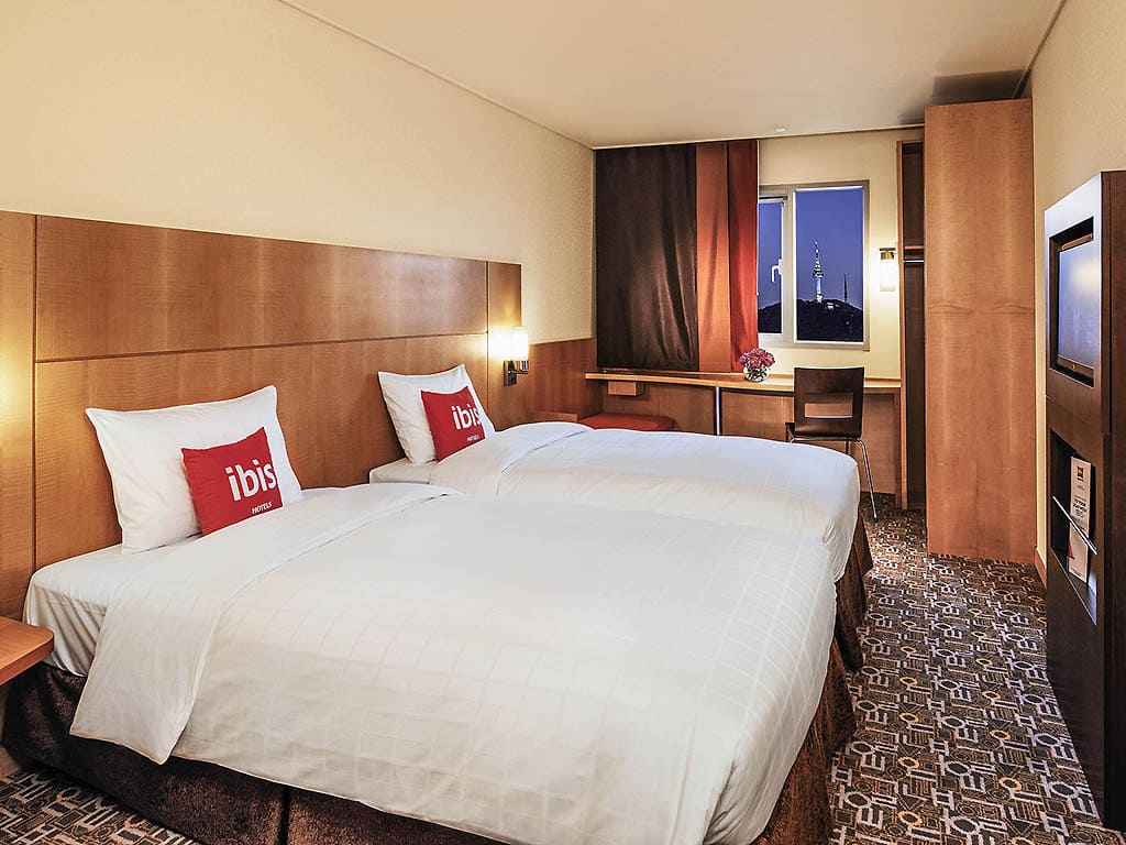 A modern hotel room with two twin beds, white linens, red ibis pillows, wooden furniture, a desk with a chair by the window, and a city view at dusk visible outside.