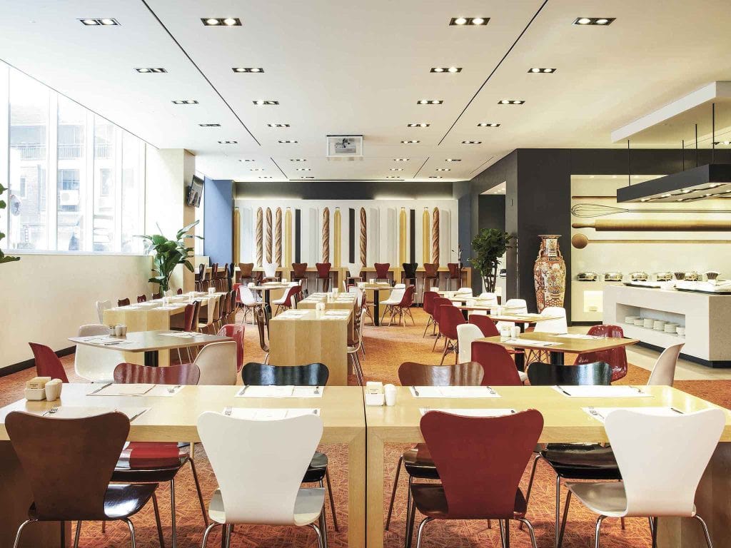 A modern, spacious dining area with multiple tables and chairs, featuring both red and white seating. Large windows let in natural light. The room includes contemporary decor and a buffet area on the right side.
