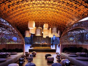 A luxurious lounge with intricate wooden lattice ceiling, round hanging lanterns, low seating, and cushions, creating a cozy, elegant atmosphere with natural light filtering in. A bar area is visible in the background.