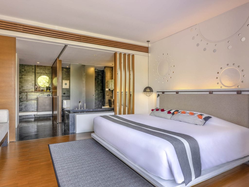 Modern hotel room with a large bed featuring white linens and gray accents, a striped decorative pillow, wooden floors, an area rug, and an open-plan bathroom with a tub and partitioned wooden slats.