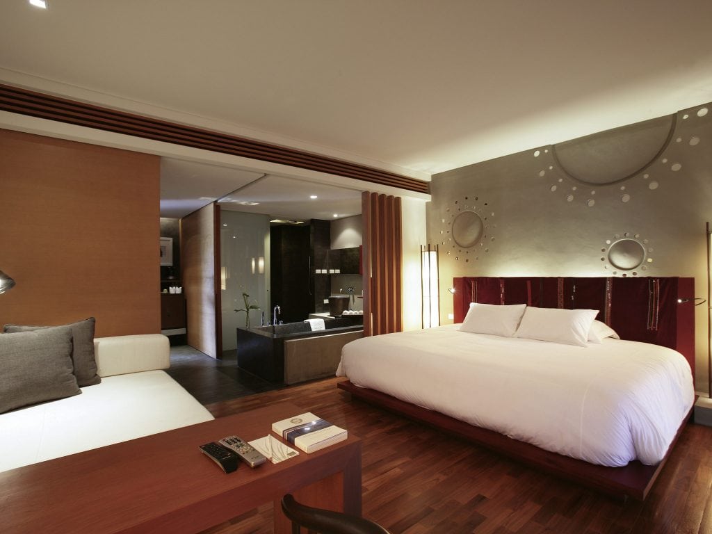 Modern hotel room with a large bed, stylish headboard, wooden floors, desk with books and remote, a white couch, and an open bathroom area with a tub and contemporary decor. Warm lighting creates a cozy atmosphere.