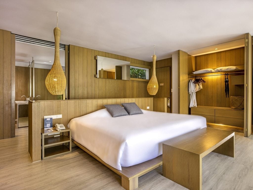 Modern hotel room with wooden walls and floor, a large bed with white linens and gray pillows, bedside tables, pendant lights, open closet with robes, and a bench at the foot of the bed.