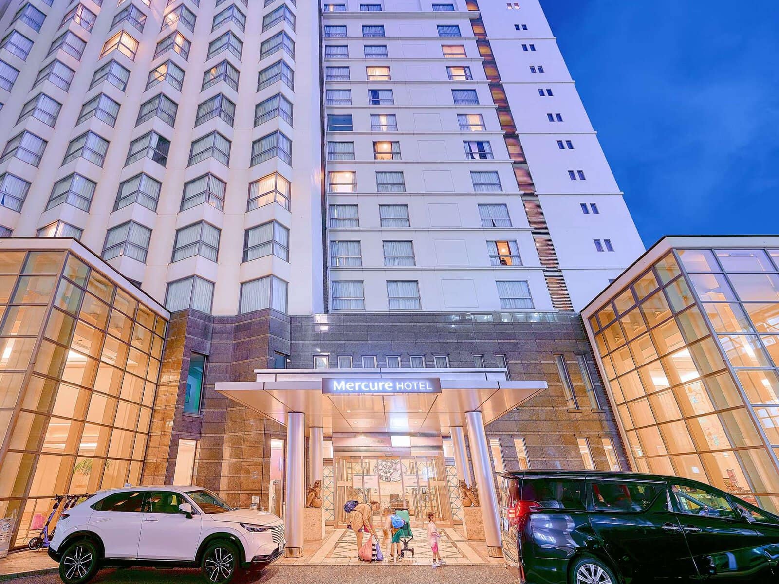 The entrance of a tall, modern hotel labeled “Mercure Hotel” at dusk, with glass panels, bright lighting, and parked cars on either side of the entrance. Three people are walking toward the doors.