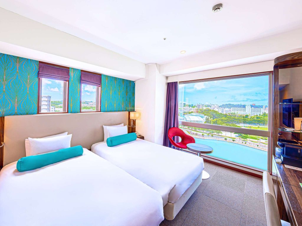 Modern hotel room with two twin beds, turquoise accents, a large window offering a city and water view, a red chair with a small table, patterned wallpaper, and bright natural lighting.