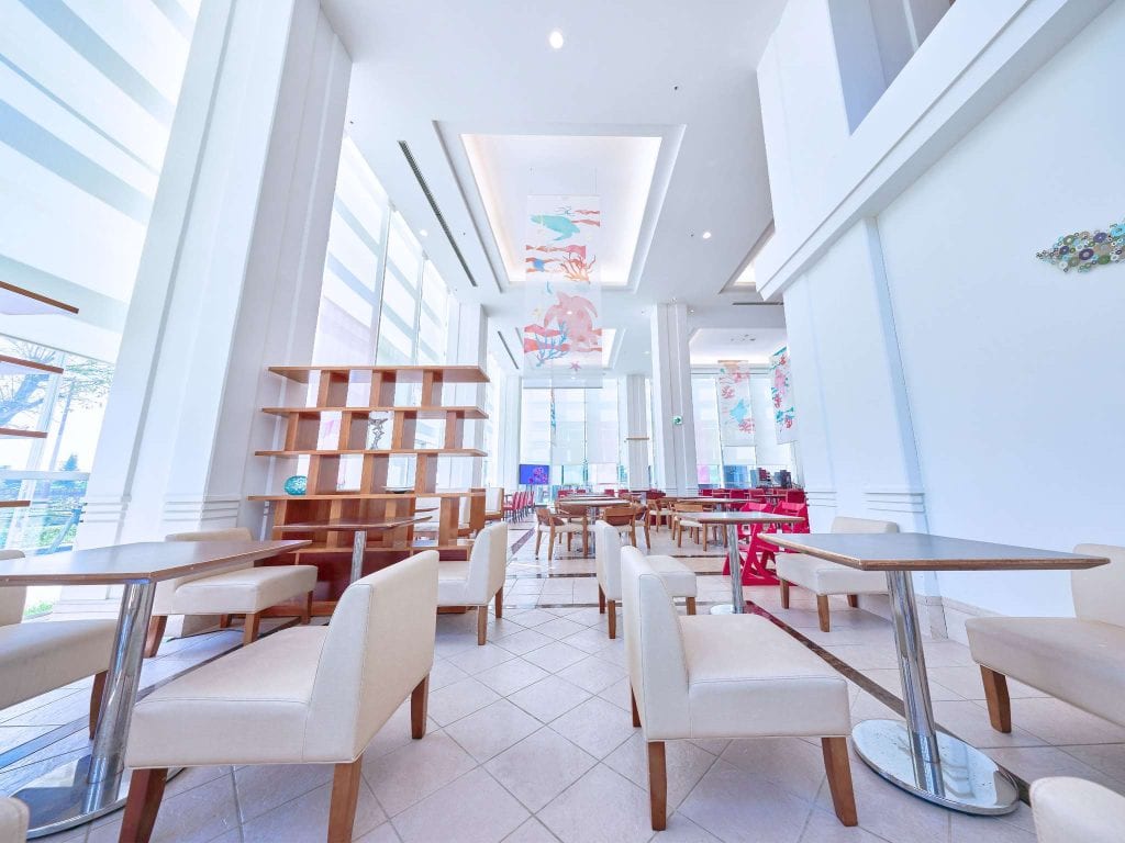 A bright, modern café with high ceilings, large windows, white walls, light wood furniture, and beige chairs. Colorful artwork decorates the walls and ceiling. The space is open and airy with natural light.