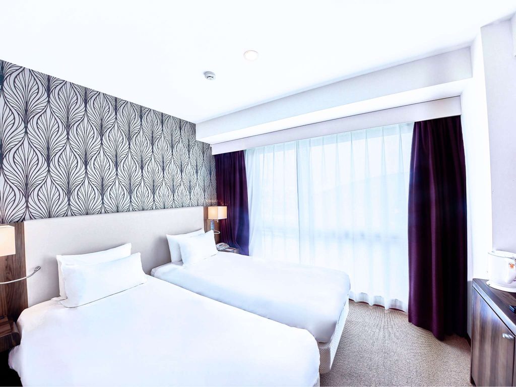 A bright hotel room with two single beds, patterned wallpaper, bedside lamps, dark curtains, large window with sheer white drapes, and a small cabinet in the corner.