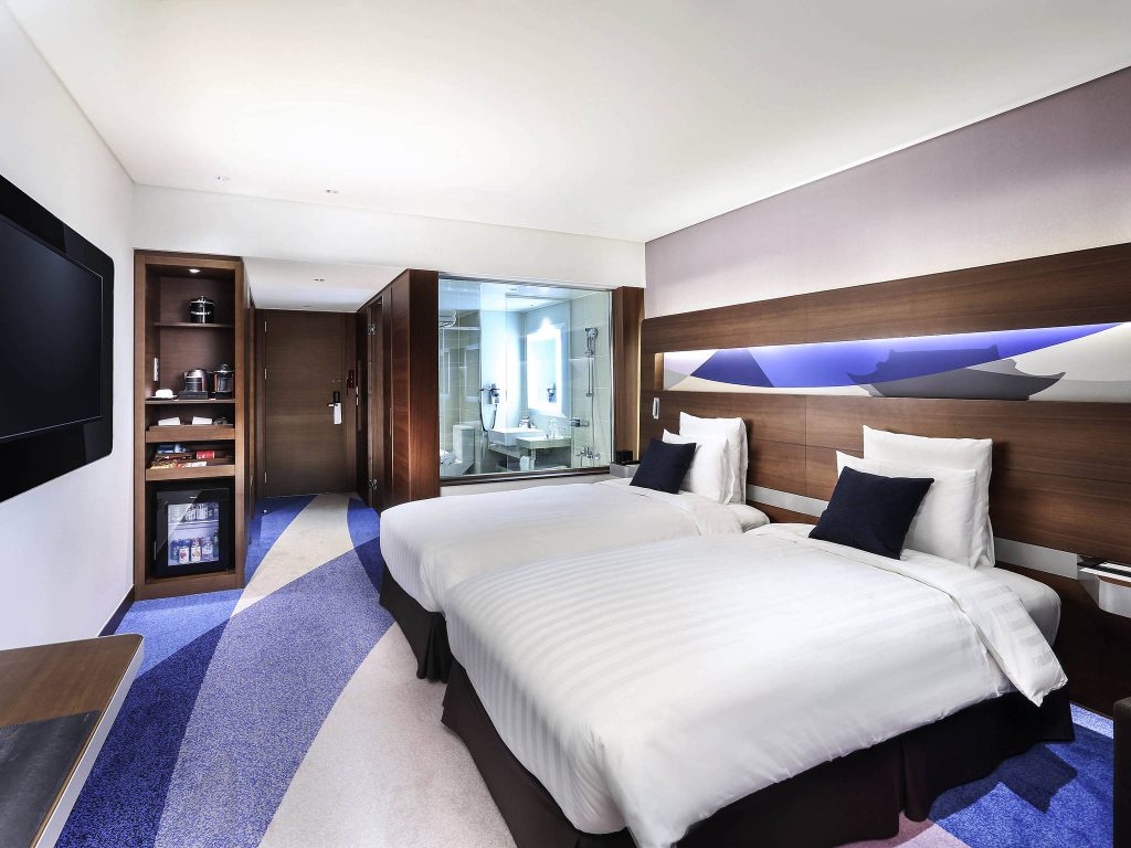 Modern hotel room with two twin beds, white linens, dark wood accents, a wall-mounted TV, and an open view of a bathroom behind a glass partition. The floor features a blue and purple wave pattern.