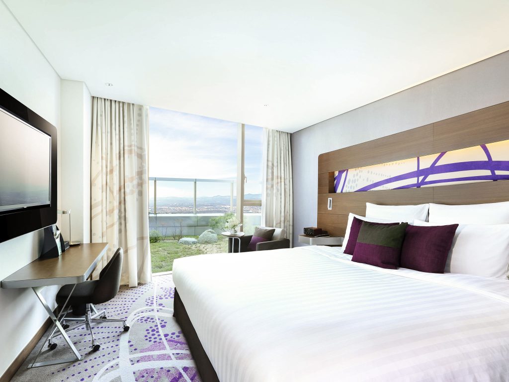 Modern hotel room with a large bed, purple accents, a desk with a chair, flat-screen TV, and floor-to-ceiling windows offering a view of a city and water outside.