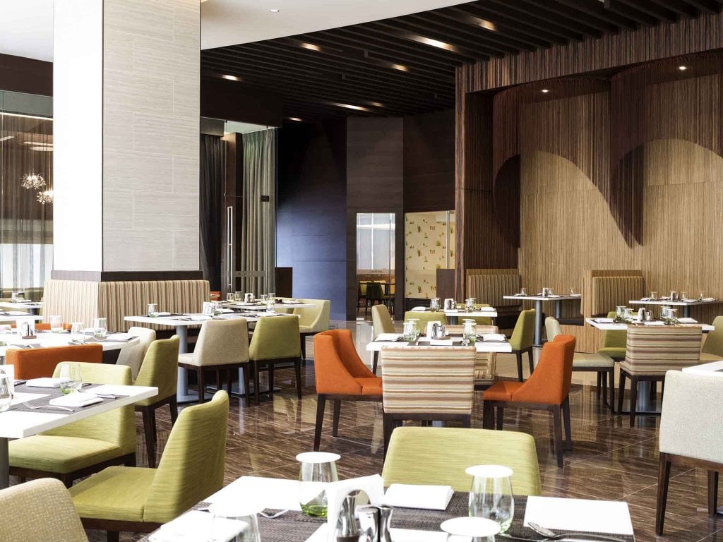 A modern restaurant dining area with tables set for service, featuring green and orange cushioned chairs, striped booths, and wooden accents. The space is well-lit with natural and artificial light.