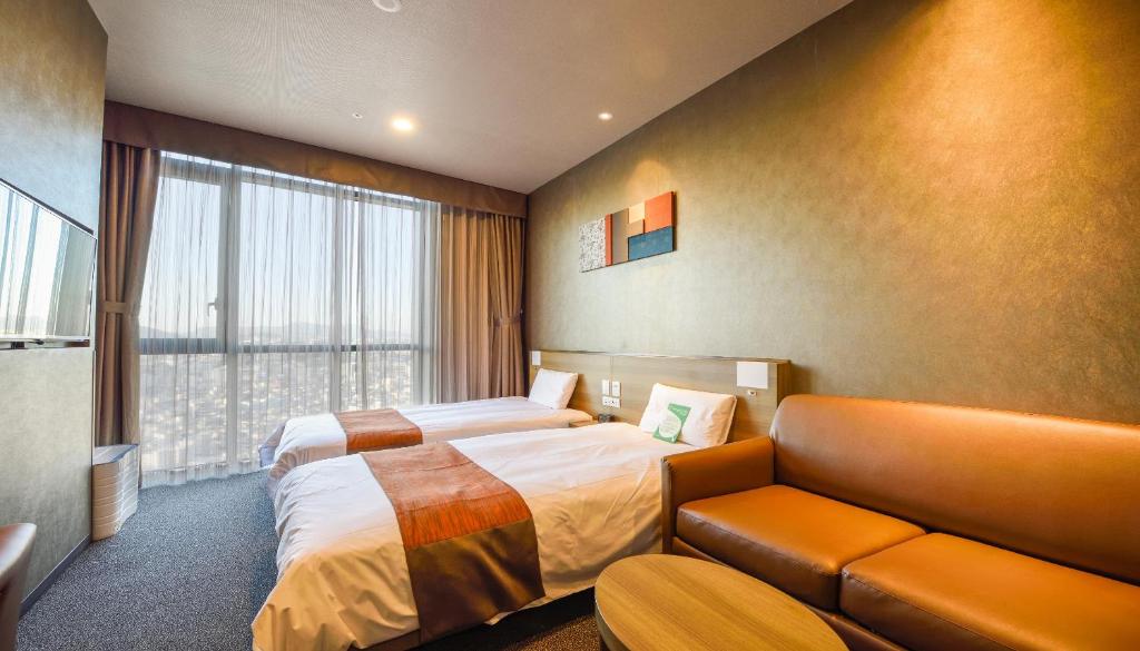 A modern hotel room with two single beds, a brown sofa, a small round table, and a large window with sheer curtains letting in daylight. The room features warm lighting and minimalist decor.