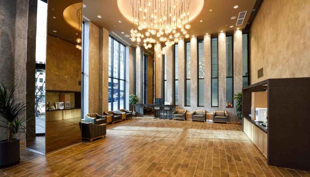 Spacious, modern hotel lobby with tall windows, several cozy armchairs, a row of vertical mirrors, a reception desk, and a ceiling adorned with numerous hanging round lights.