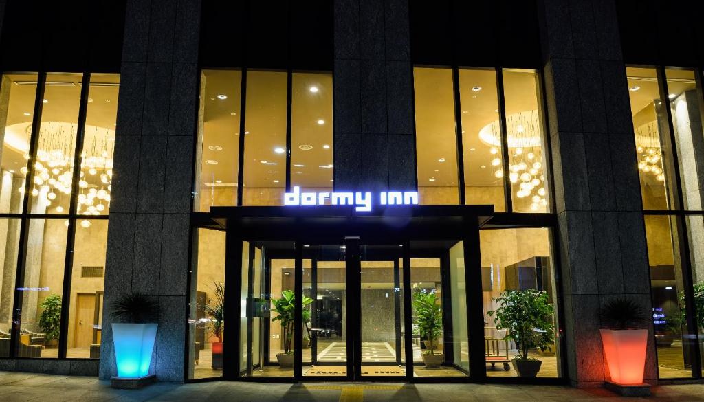 The entrance of a modern hotel at night, with tall glass doors, lit interior, decorative plants, and a glowing dormy inn sign above the doorway.