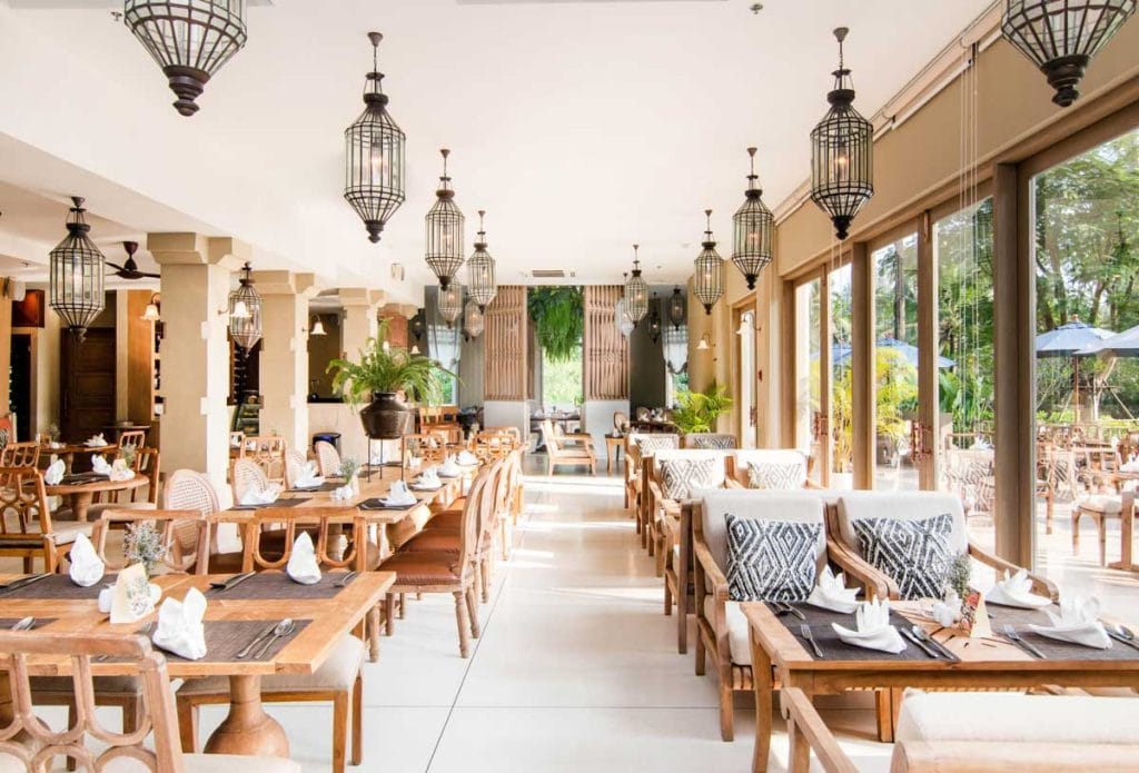 A bright, spacious restaurant with wooden tables and chairs, decorative lanterns hanging from the ceiling, large windows letting in natural light, and greenery visible outside. Tables are set with napkins and glassware.