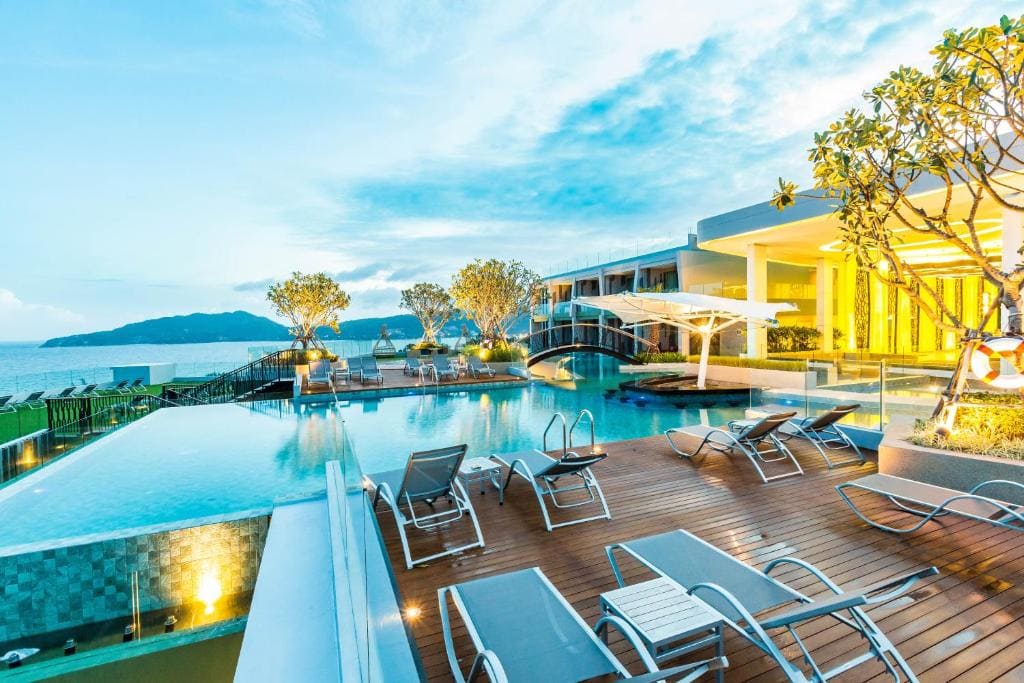 Modern beachfront resort with a large outdoor pool, sun loungers on a wooden deck, and a view of mountains across the water at sunset. The building is brightly lit and surrounded by trees.
