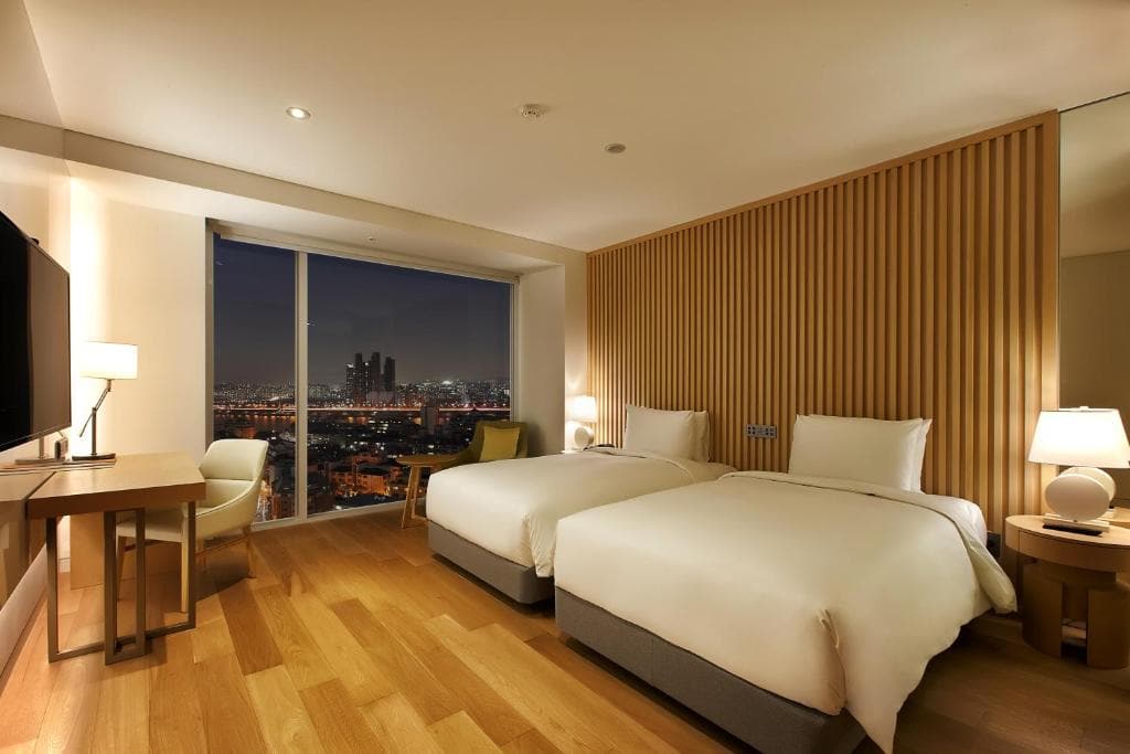 Modern hotel room with two single beds, wooden floors, a desk, lamps, and a large window offering a nighttime city view with illuminated buildings outside. The room has a warm, inviting ambiance.