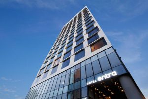 Tall modern high-rise hotel building photographed from below, with a glass and metal facade. The sign near the entrance reads HOTEL entra. Clear blue sky in the background.