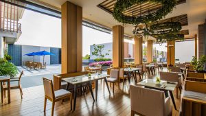 Modern open-air restaurant with wooden floors and furniture, greenery decor hanging from the ceiling, and outdoor seating visible with blue umbrellas on a sunny day.