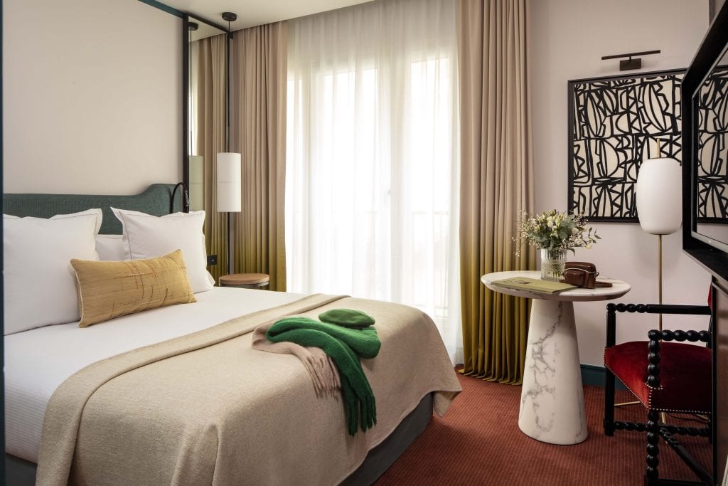 A stylish hotel room with a neatly made bed, beige bedding, green throw, and decorative pillows. A round marble table with flowers, modern artwork, and a window with long curtains complete the cozy, modern decor.