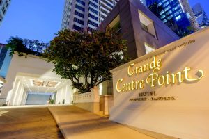Entrance of Grande Centre Point Hotel in Bangkok at dusk, with illuminated signage in the foreground and tall buildings and greenery in the background.