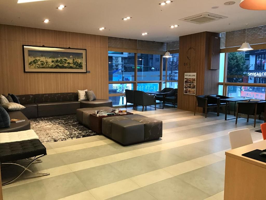 A modern, spacious lounge with a large sectional sofa, ottomans, armchairs, and tables. Large windows let in natural light. Wooden walls, a rug, and wall art add warmth to the room. City buildings are visible outside.
