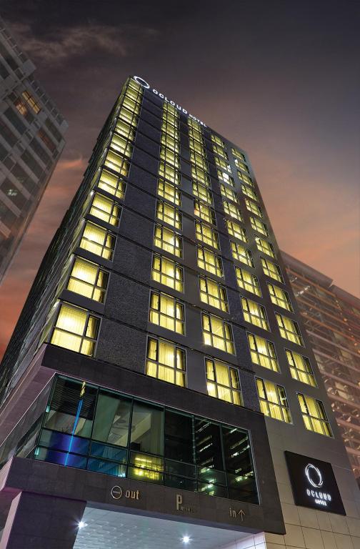 A tall, modern hotel building illuminated at night, with numerous lit windows and a well-lit entrance. The sky is dusky, and nearby buildings are partially visible. The hotels name, Oakland Hotel, is displayed at the top.