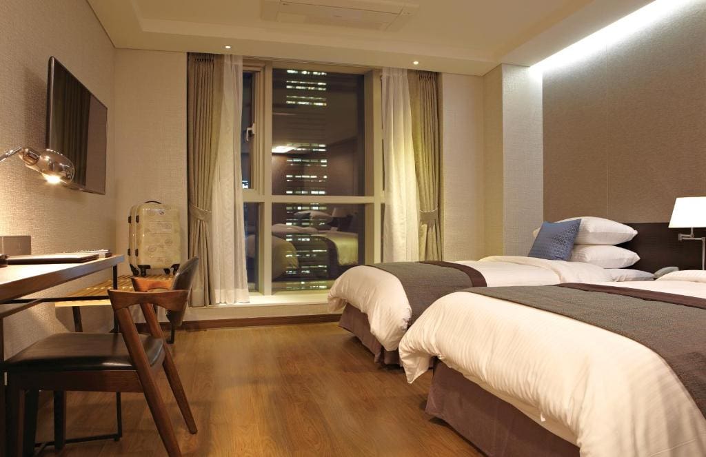 A modern hotel room with two neatly made twin beds, a desk with a lamp and chair, a mounted TV, and a large window showing city buildings at night. The room has wooden flooring and neutral-colored walls.