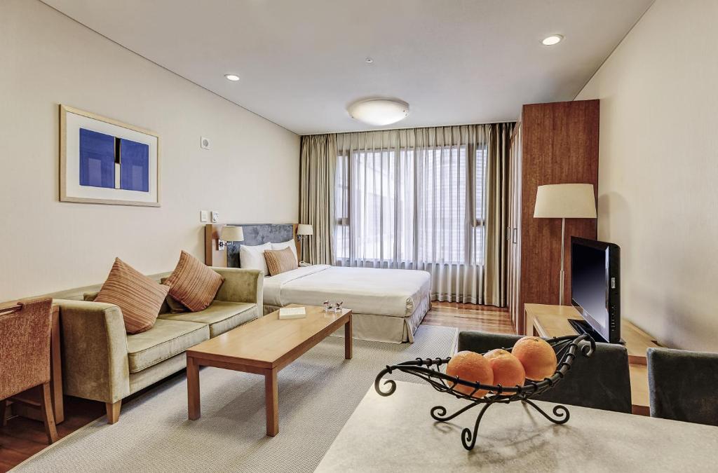 Modern hotel room with a double bed, sofa, coffee table, TV, and a large window with sheer curtains. A bowl of oranges sits on a table in the foreground. The space is cozy with neutral colors and soft lighting.