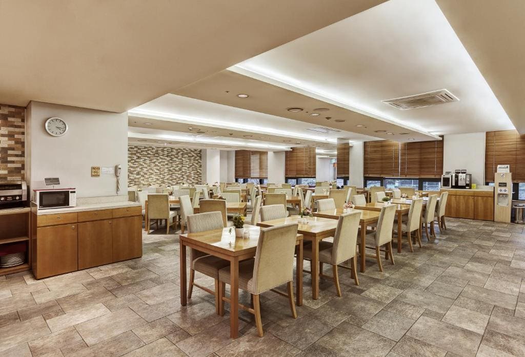 A modern, spacious dining area with wooden tables and cream chairs neatly arranged. The room features tiled floors, wood-paneled walls, and a buffet counter with microwave, water dispenser, and coffee machines on the left side.