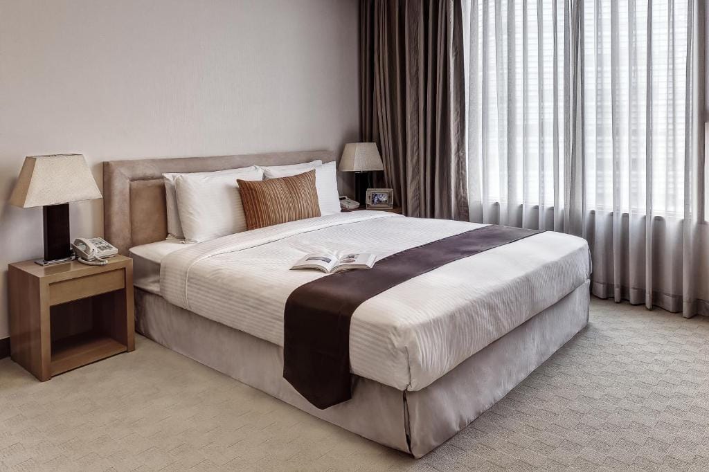 A neatly made bed with white linens, brown accent pillows and runner, and an open book lies on top. Bedside tables with lamps are on either side, and a large window with sheer curtains lets in natural light.