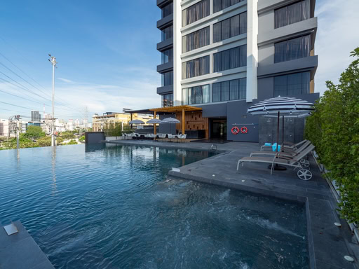 A modern hotel pool area with lounge chairs, umbrellas, and a shaded seating area. The pool has clear water and overlooks a cityscape with buildings under a blue sky.