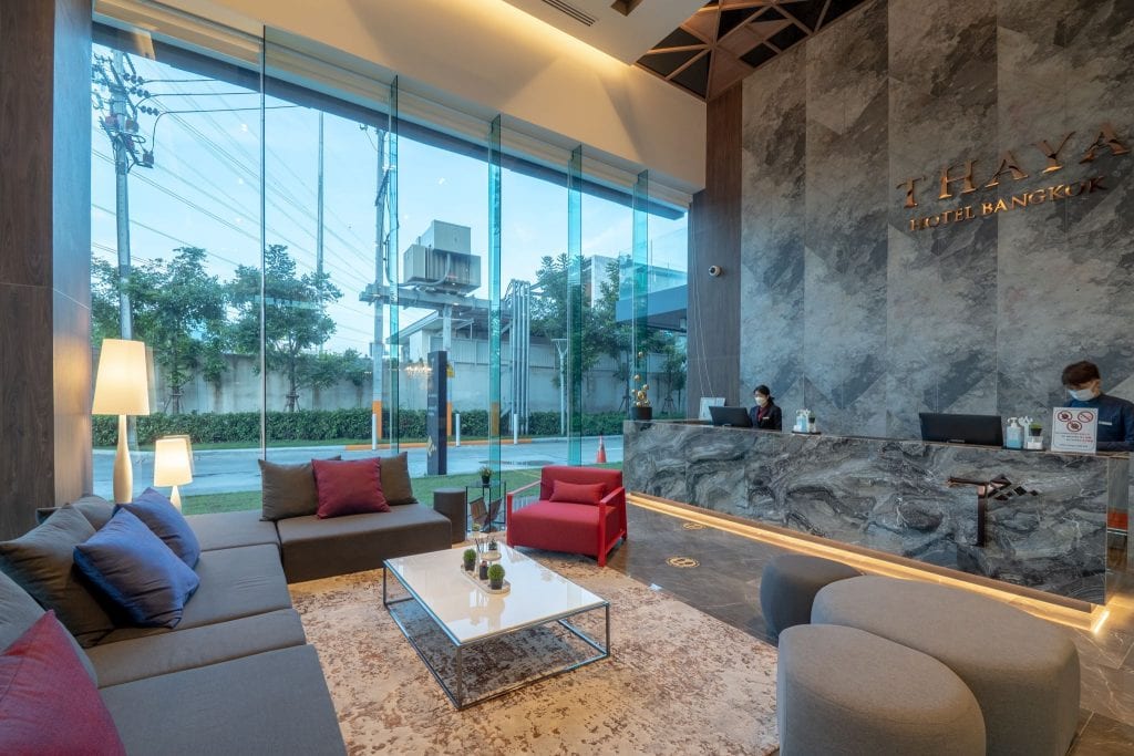 Modern hotel lobby with large floor-to-ceiling windows, gray sofas, a red armchair, a coffee table, and a marble reception desk where a staff member is assisting a guest. The setting appears bright and welcoming.