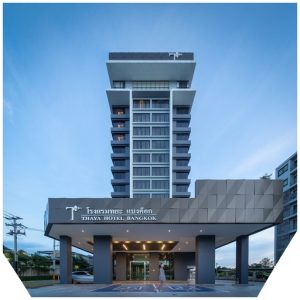 A modern high-rise hotel with a gray facade, large windows, and a covered entrance. The sign reads THAYA HOTEL BANGKOK in English and Thai. A person walks near the entrance under a blue sky.