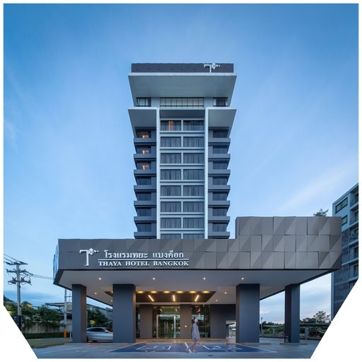 A modern high-rise hotel with a gray facade, large windows, and a covered entrance. The sign reads THAYA HOTEL BANGKOK in English and Thai. A person walks near the entrance under a blue sky.