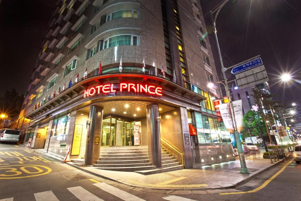 A brightly lit hotel entrance at night displays a red neon sign reading HOTEL PRINCE. The building has multiple flags above the entrance and is located on a city street with traffic signs and streetlights.