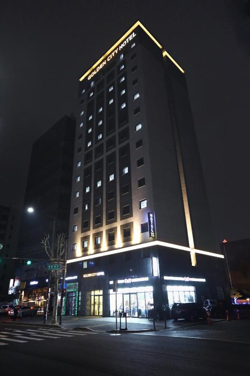 A tall hotel building with illuminated Golden City Hotel signs at night, featuring many lit windows and a bright ground-floor entrance along a city street with cars and trees visible.
