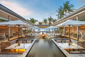 Upscale outdoor restaurant with sunken seating areas around a reflective pool, white umbrellas, elegant table settings, and palm trees; ocean and sunset visible in the background.