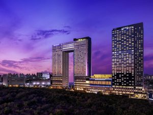 A modern cityscape at dusk featuring two illuminated high-rise hotels; one is rectangular with a large central cutout, and the other is a tall, slim building. The sky is purple and pink, and city lights glow in the background.