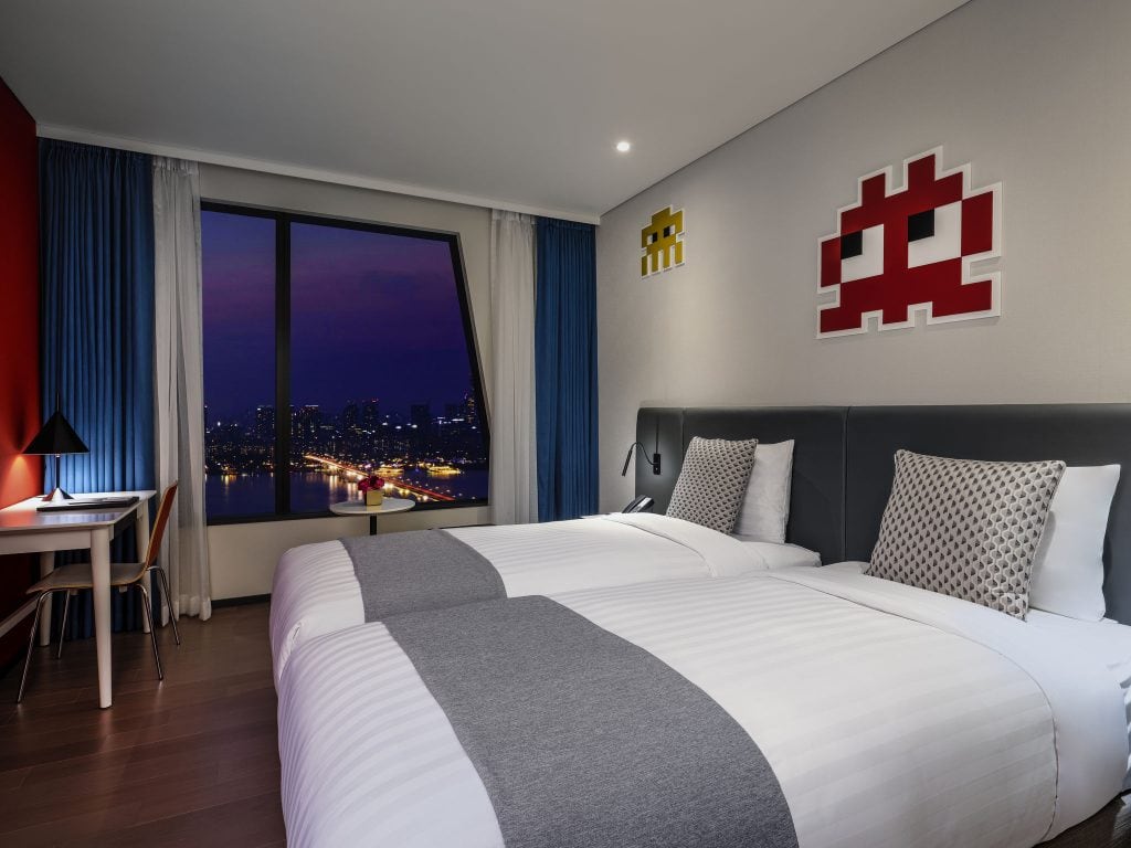 Modern hotel room with two single beds, gray and white bedding, geometric pillows, a desk by a large window with city night view, and retro video game art on the wall above the beds.