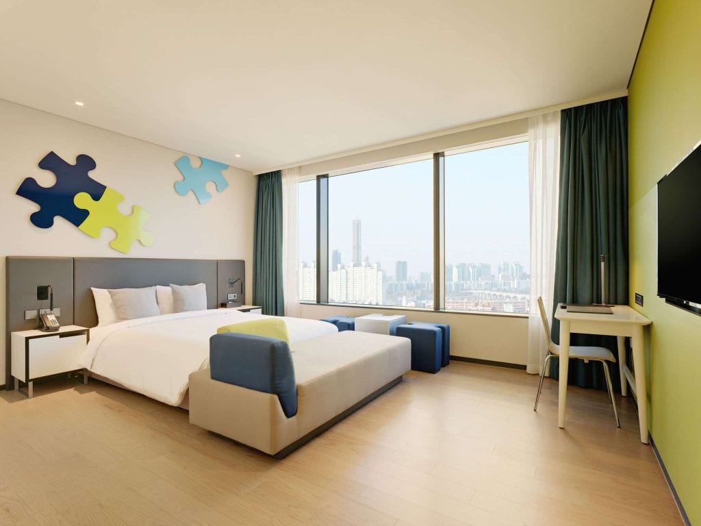 Modern hotel room with a large bed, blue and lime décor, wall art shaped like puzzle pieces, a bench, desk, TV, and large windows revealing a city skyline view. Light wood flooring and natural light fill the space.