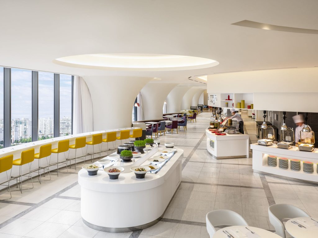 A modern, bright buffet restaurant with curved white walls, large windows, yellow barstools, and a variety of food dishes displayed on sleek white counters. Chefs prepare food in the open kitchen area.