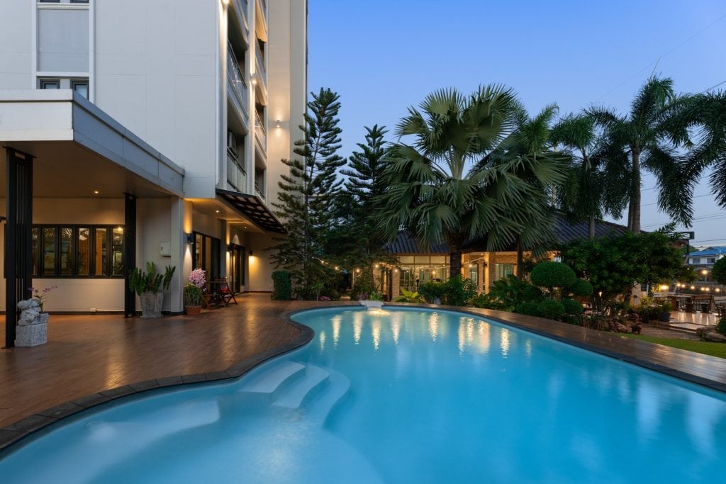 A serene outdoor pool curves alongside a modern building at dusk, surrounded by palm trees, manicured bushes, and a gazebo with warm lights, creating a relaxing resort ambiance.