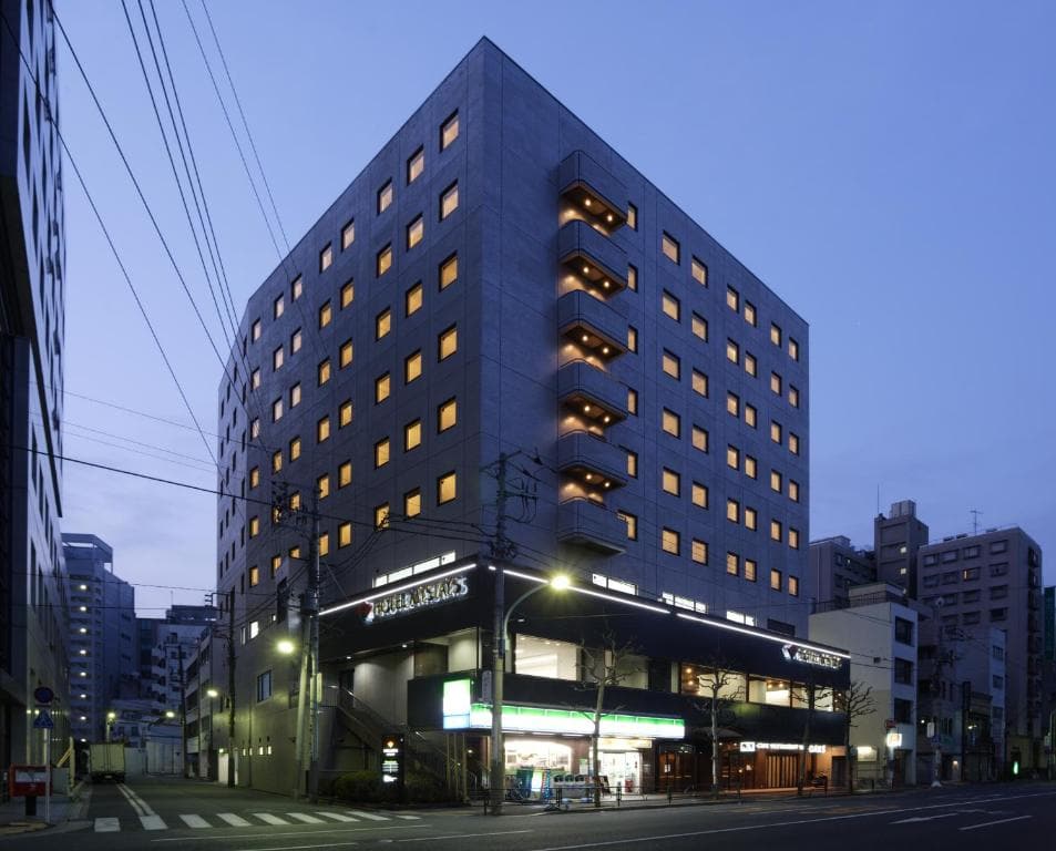 A modern, multi-story hotel building at dusk with many windows lit up. A convenience store is on the ground floor, and streetlights illuminate the sidewalk and crosswalk in an urban setting.