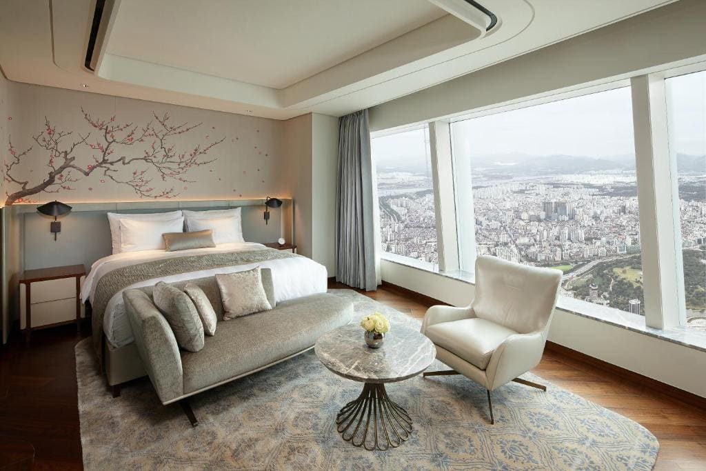 A modern hotel room with a large bed, a cushioned bench, a marble round table, a white armchair, and floor-to-ceiling windows offering a panoramic city view. The walls feature a cherry blossom mural above the bed.