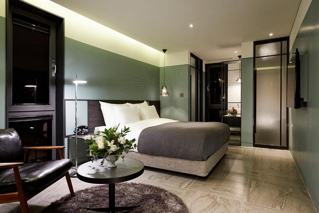 Modern hotel room with a double bed, gray bedding, green accent walls, a small round table with a flower arrangement, a black armchair, and a softly lit, minimalist design.