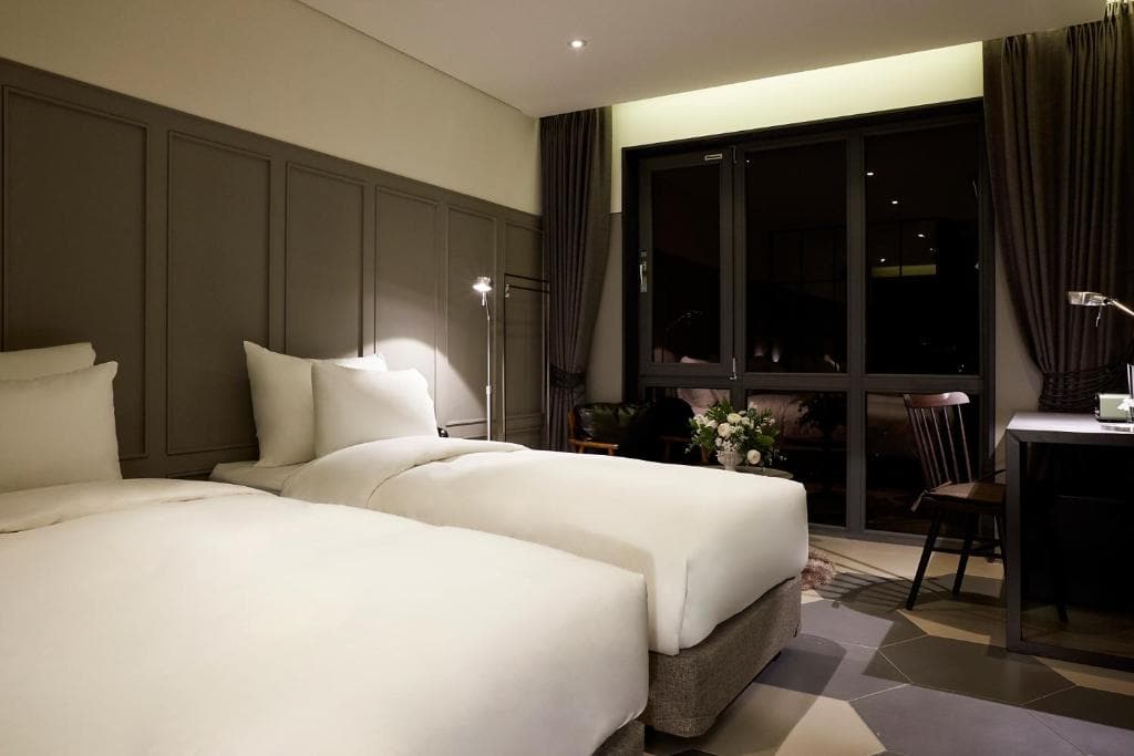 A modern hotel room with two neatly made single beds, dark paneled walls, a large window with closed curtains, a desk with a lamp and chair, and a small table with flowers. The lighting is soft and warm.