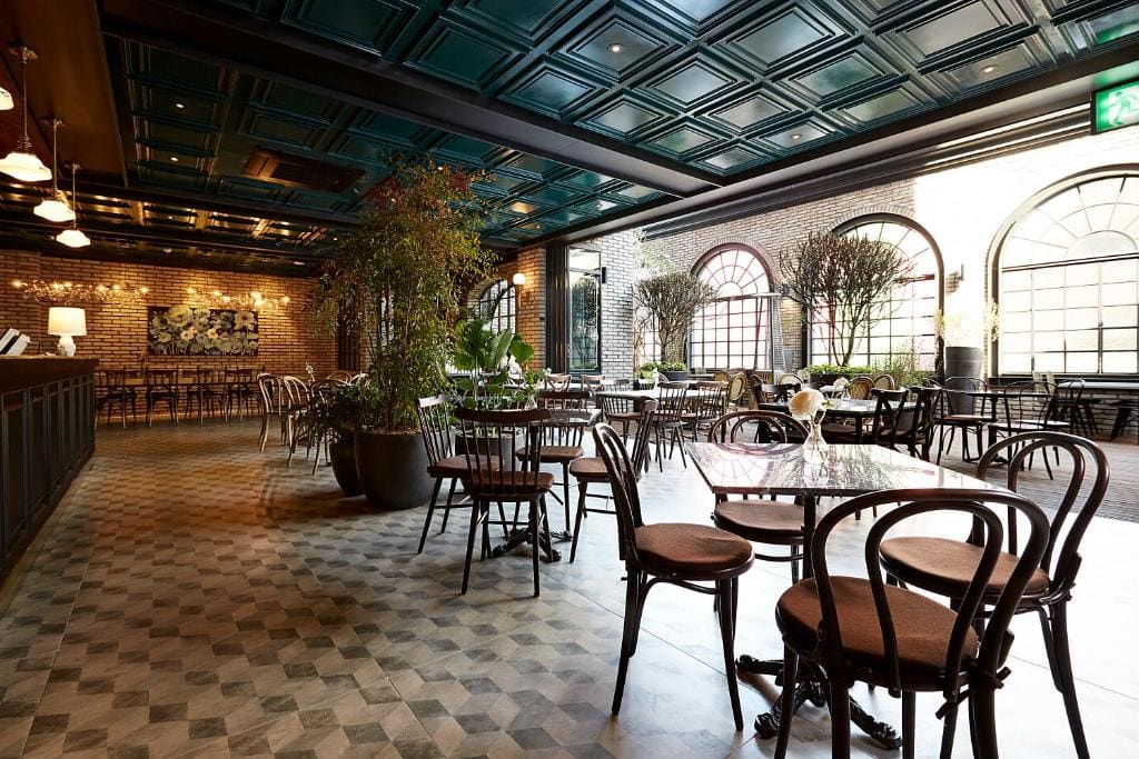 Spacious, sunlit café with large arched windows, brown chairs and tables, potted plants, a tiled floor, and a high, green coffered ceiling. The setting feels elegant and inviting.