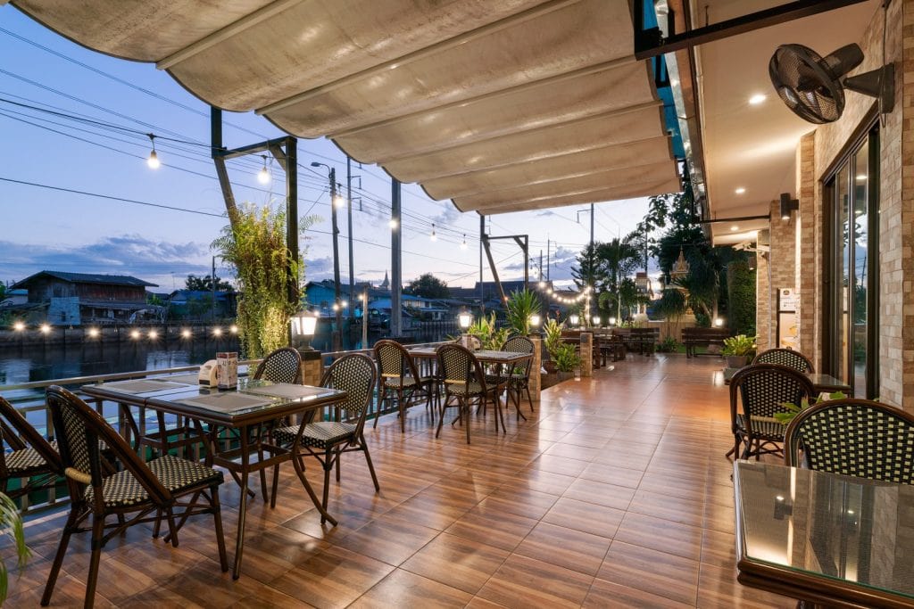 Outdoor restaurant terrace with wicker chairs and tables overlooks a river at dusk. String lights and greenery create a cozy atmosphere, while the setting sun casts a gentle light over the calm scene.
