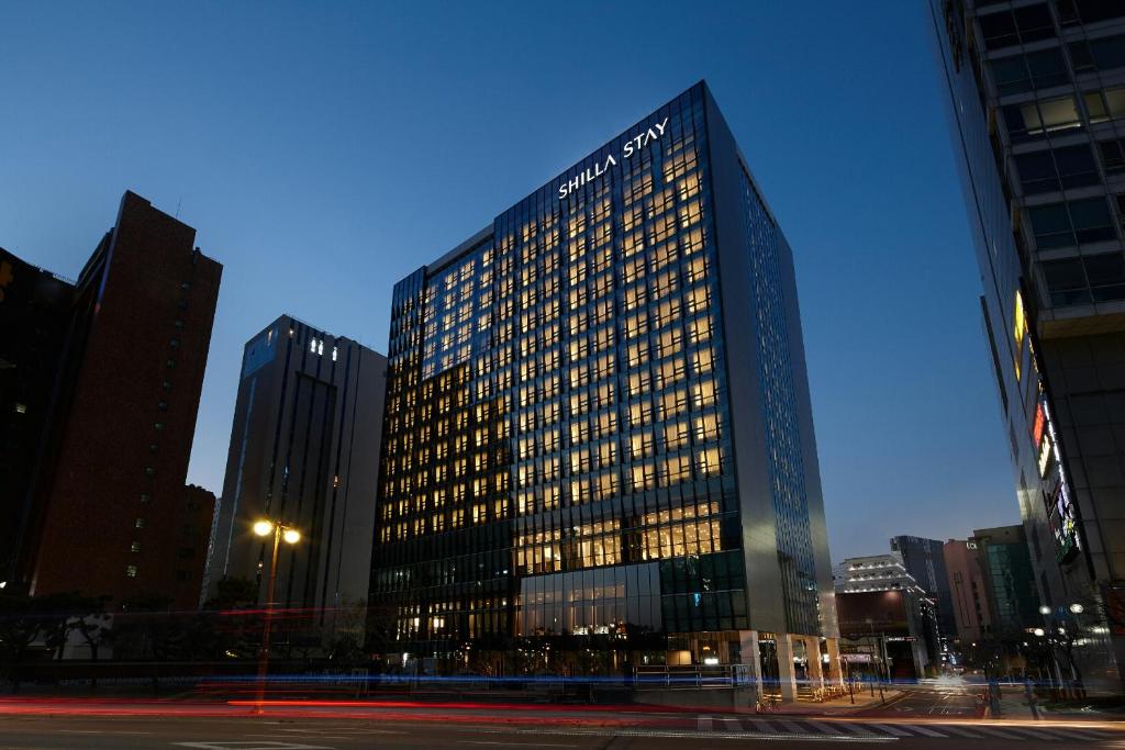 A modern high-rise hotel, Shilla Stay, is illuminated at dusk with many windows lit up. The building stands at a city intersection, surrounded by other tall structures and a clear evening sky.