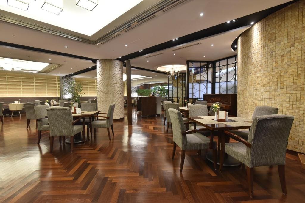 A spacious, modern restaurant dining area with wooden floors, brick accent walls, gray upholstered chairs, and square tables, softly lit by ceiling lights and decorative lamps.